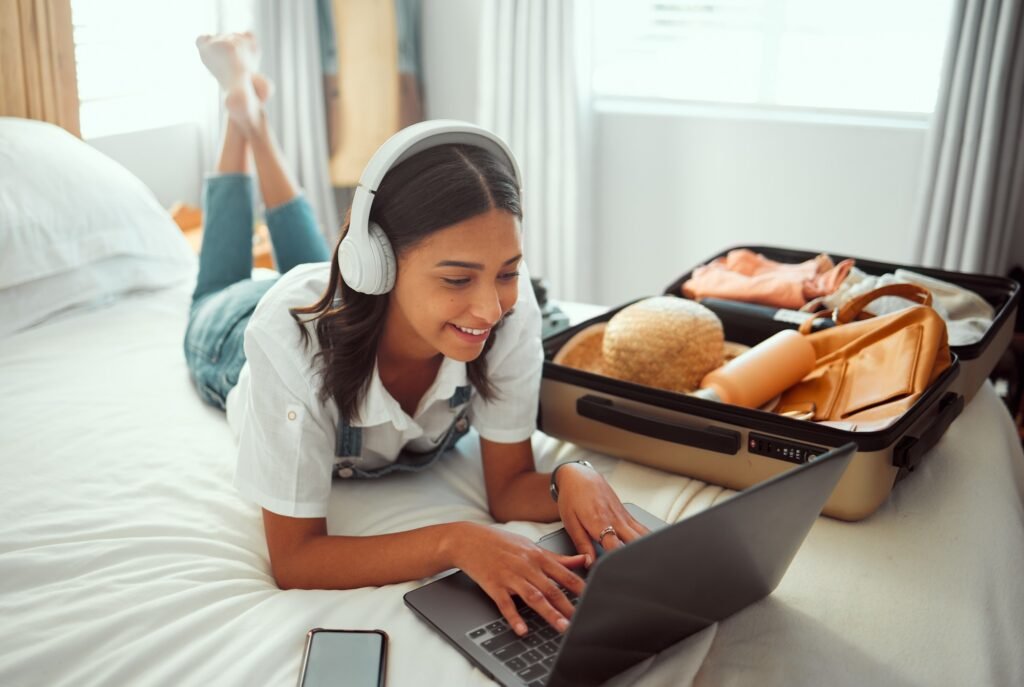 Computer travel checklist and woman packing for a vacation with headphones. Adventure, planning and female happy on bedroom with a luggage, suitcase and bag ready for a journey.